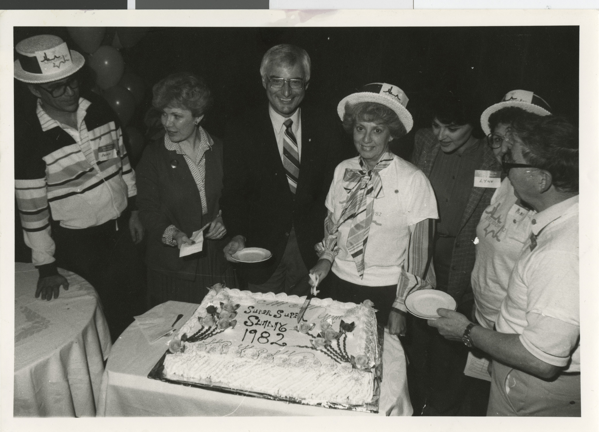 Photograph of cake cutting for Super Sunday event with Dennis Sabbath, Myrna Williams, Jim Santini, Dorothy Eisenberg (cutting cake), Lyn Rosencrantz, and Ray and Jerry Countess, 1982